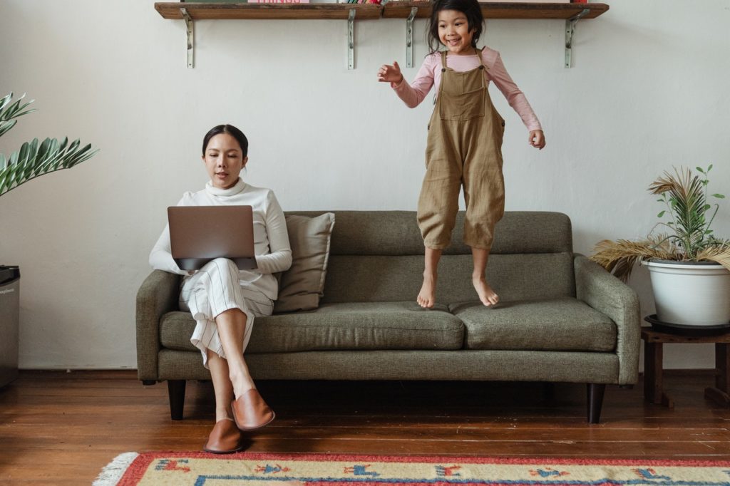 woman working on the couch with her daughter jumping next to her